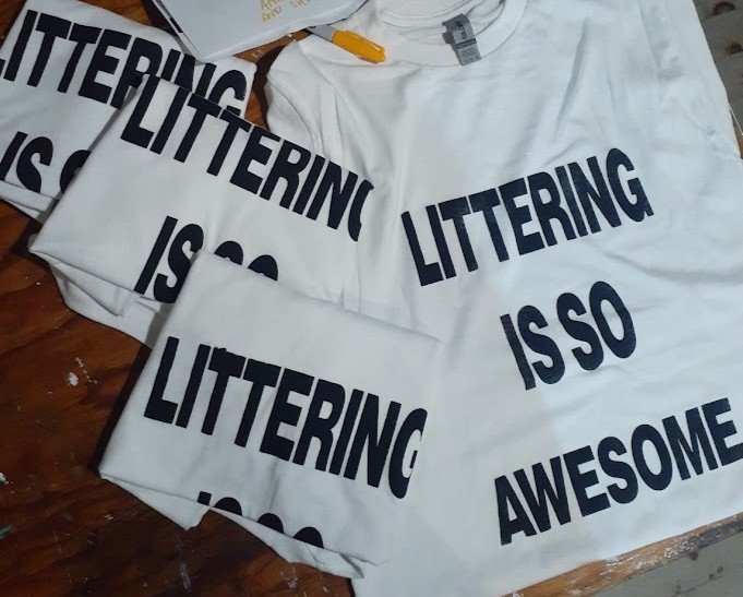 Crisis Hotline merch: white t-shirts reading "Littering is so awesome" in blue block letters